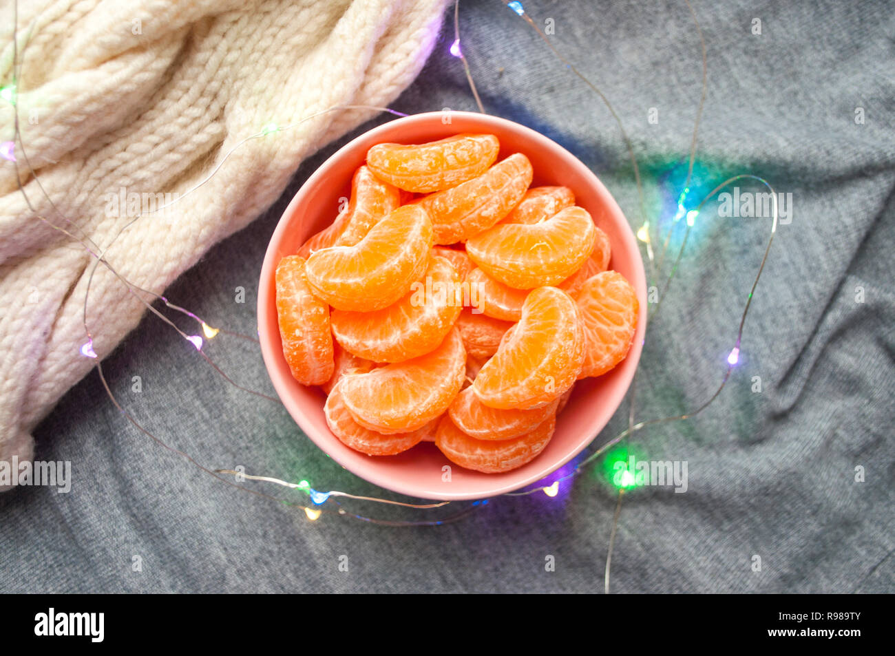 Flat lay of pink bowl full of peeled sweet tangerines on grey and white knitted background with garland lights. Stock Photo