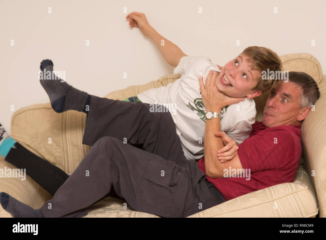 grandfather or father fighting, play-fighting, wrestling, playing rough, with son or grandson, youth, twelve-years-old,  on sofa settee, having fun. Stock Photo