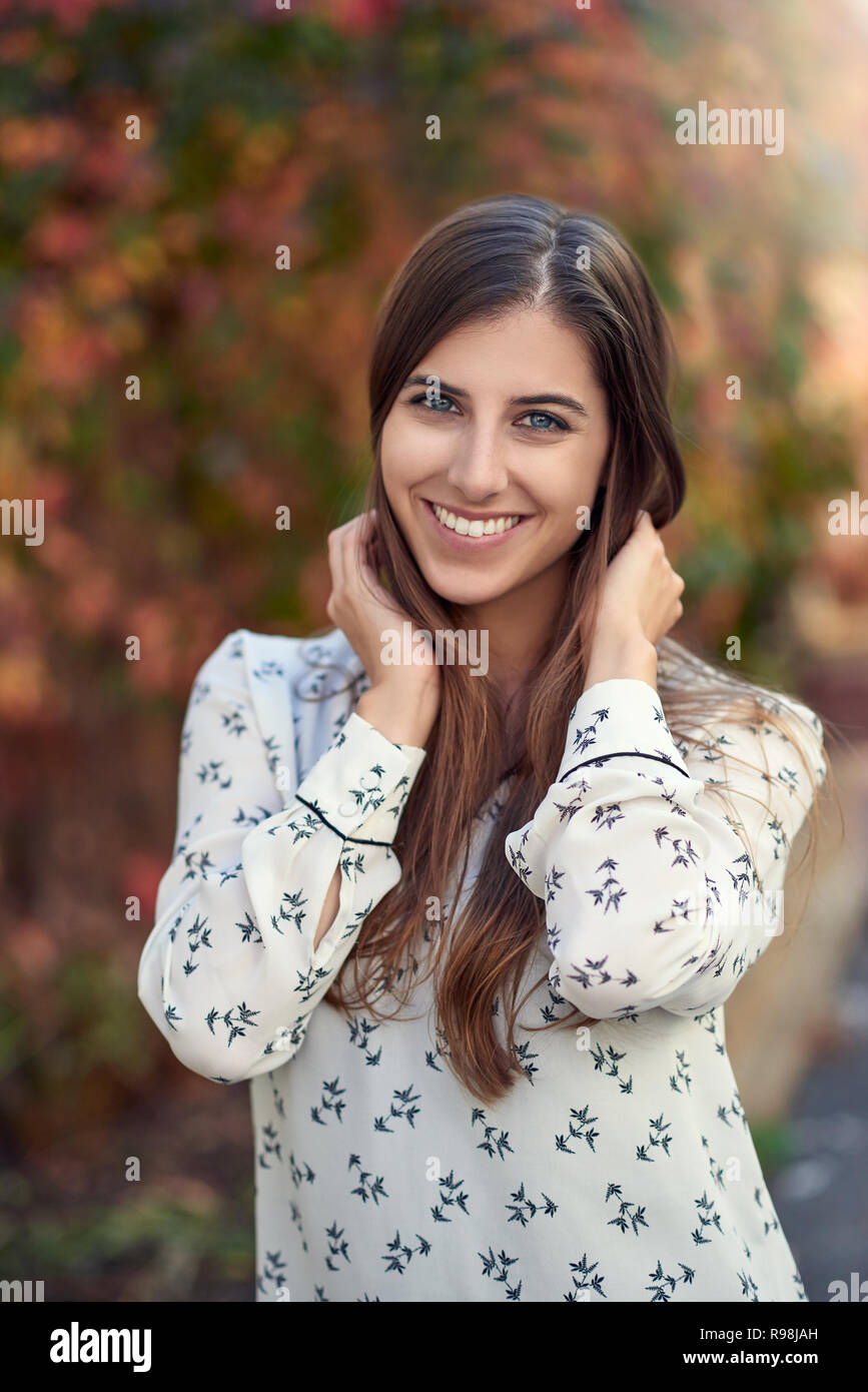 Attractive young woman in a colorful fall or autumn street standing sideways to the camera with her hand to her cheek smiling at the camera Stock Photo