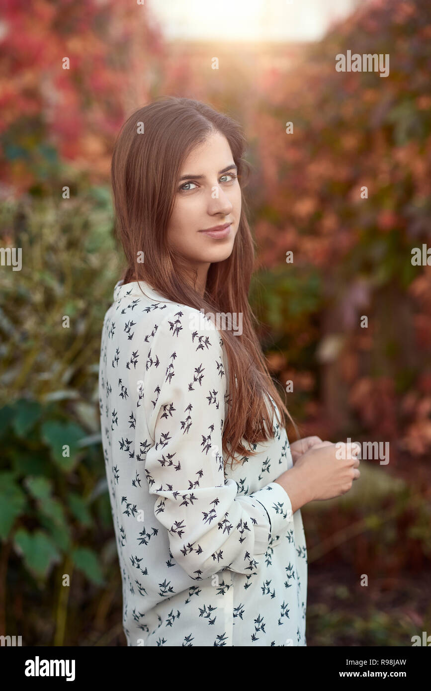 Attractive young woman in a colorful fall or autumn street standing sideways to the camera with her hand to her cheek smiling at the camera Stock Photo