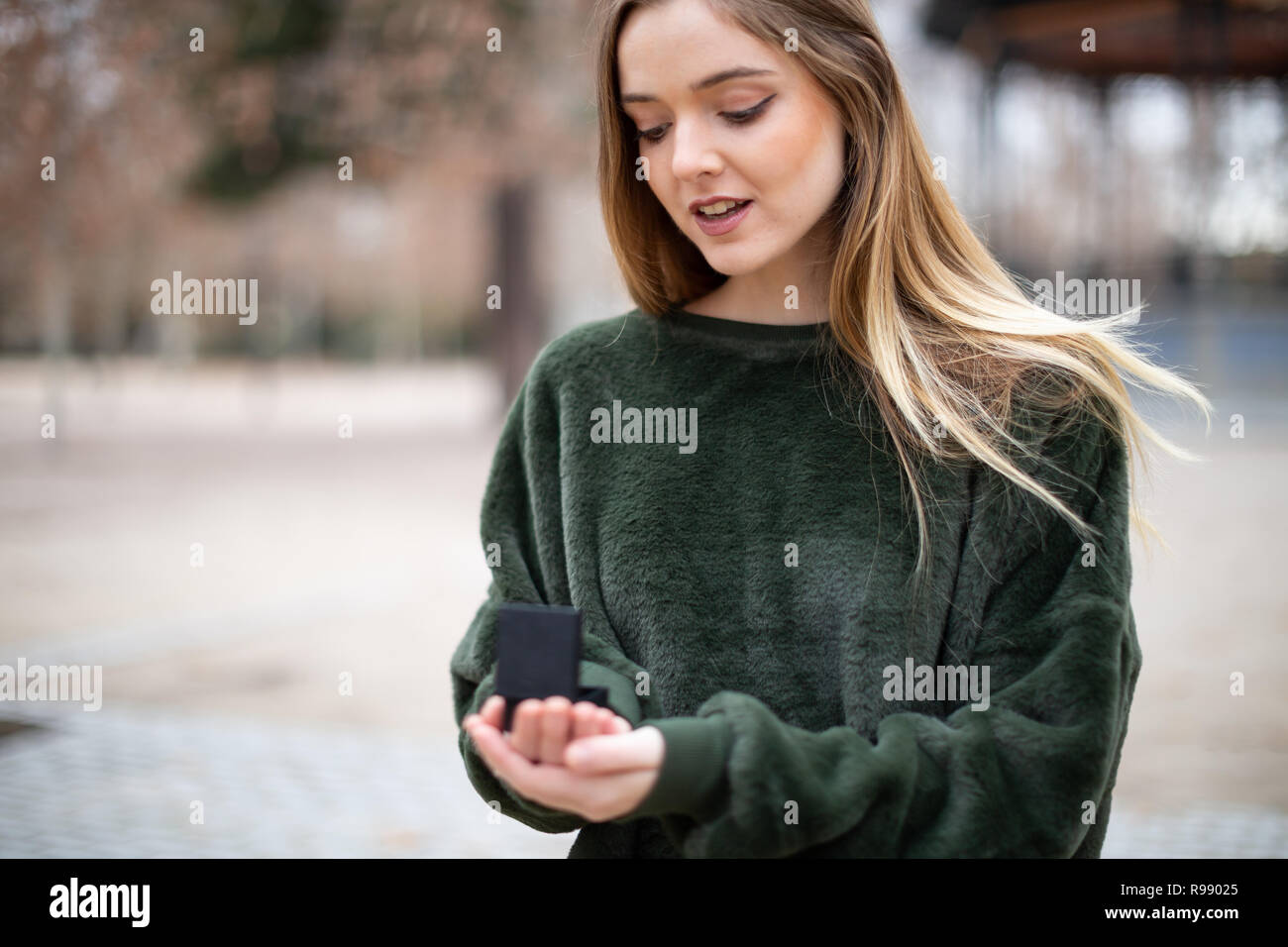 Beautiful young lady in stylish outfit looking at wedding ring in box while standing on blurred Stock Photo