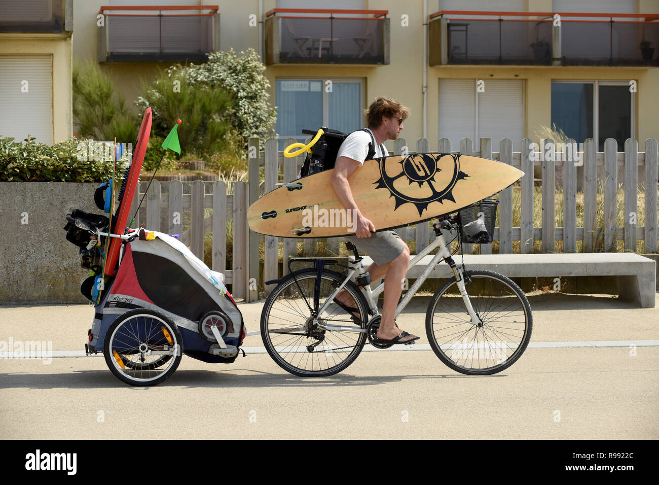 Surfer riding bike with surf board and towing child in trailer at Wissant in northern France Stock Photo