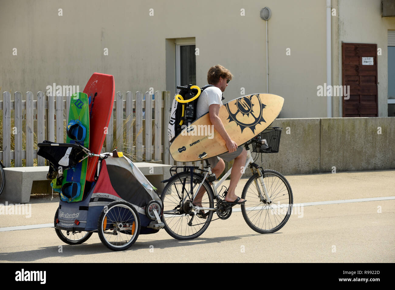 Surfer riding bike with surf board and towing child in trailer at Wissant in northern France Stock Photo