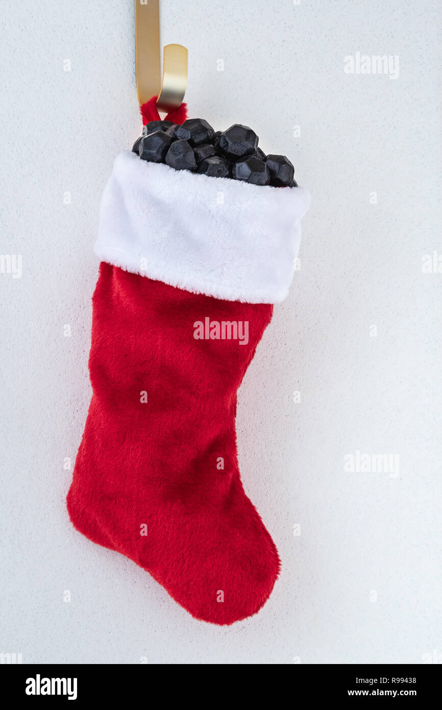 Traditional red and white plush Christmas stocking stuffed with coal shaped candy on a gold hook against a white background Stock Photo