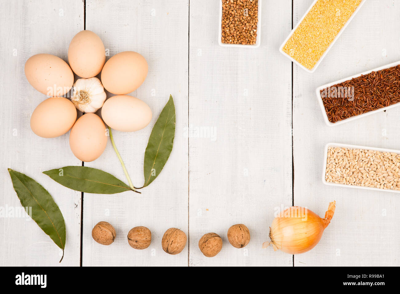 Various groats in bowls, eggs, bay leaf laid out in the shape of a flower on white wooden table Stock Photo