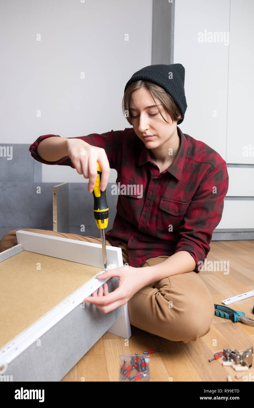 Woman assembling a chest drawer for new bedroom furniture. Female in casual shirt and knit hat does typical 'men' carpenter work at home Stock Photo