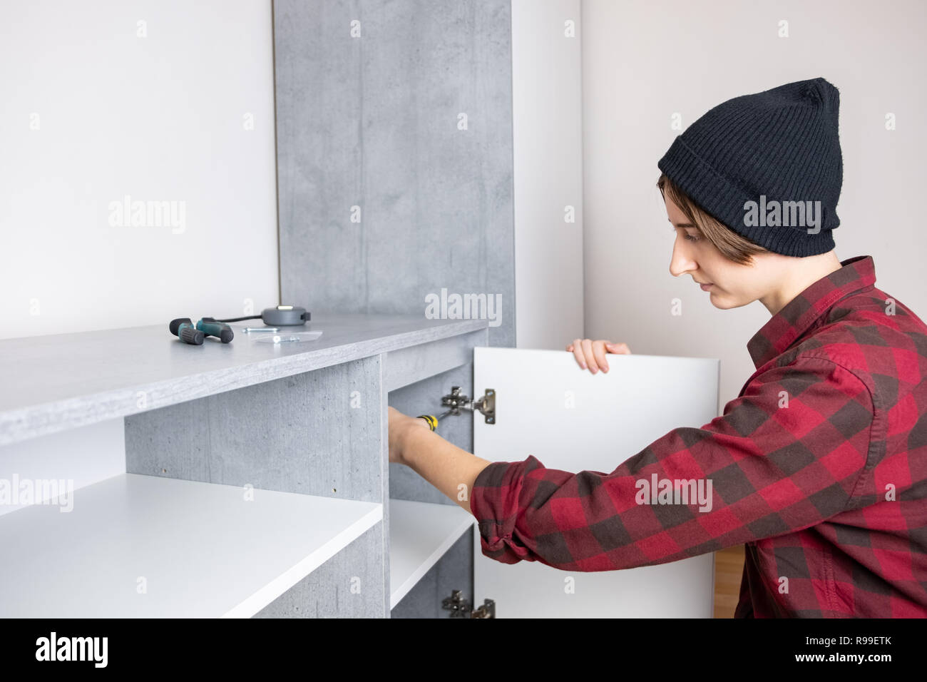 Woman adjusting furniture door in a new living room cabinet. Female in casual shirt and knit hat does 'men' carpenter work at home Stock Photo