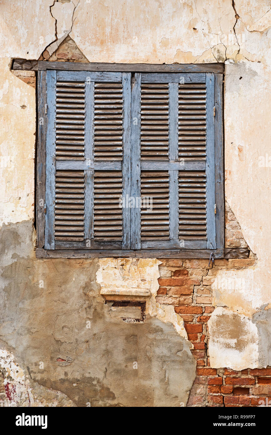 old blue shuttered window in cracked and peeling plaster wall over bricks Stock Photo