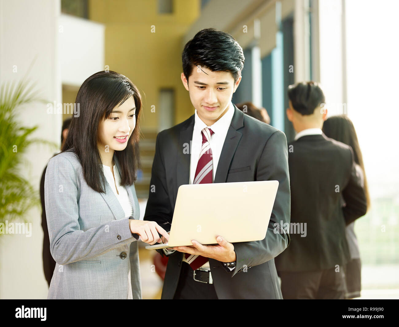 young asian businessman and businesswoman working together in office using laptop computer, colleagues talking in background. Stock Photo