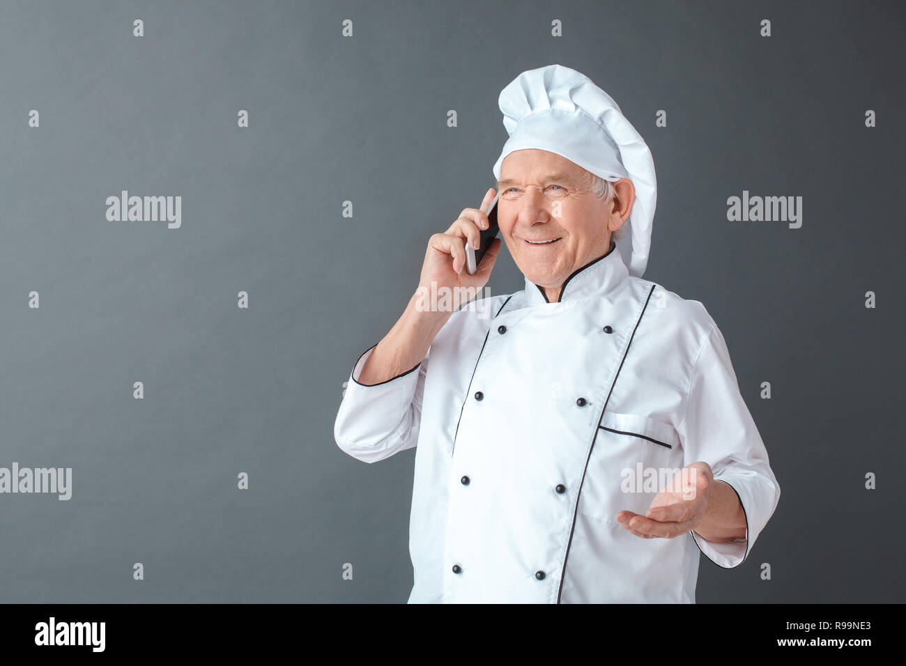 Senior male chef studio standing isolated on gray wall answering phone ...