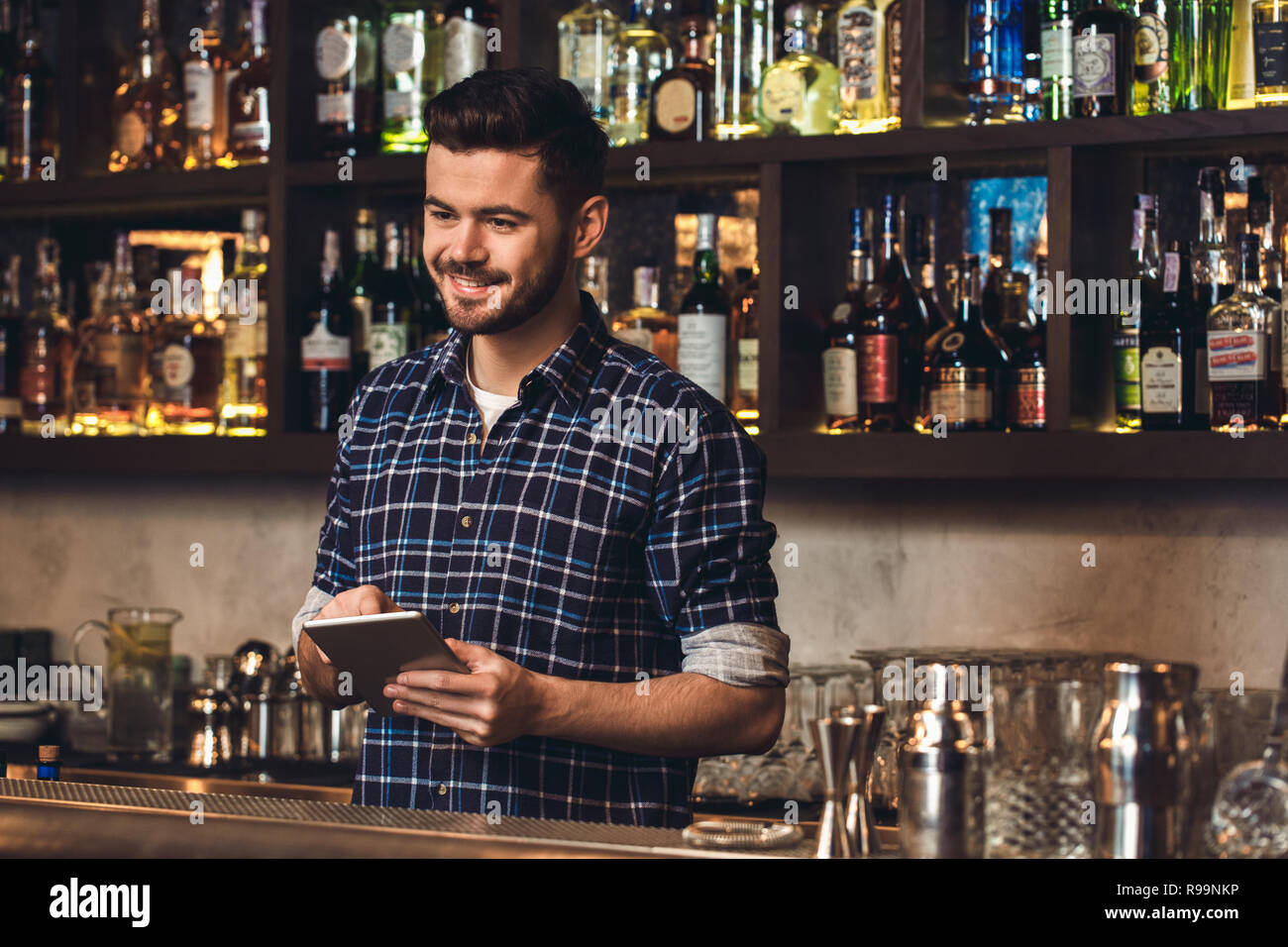 Young barman standing at bar counter placing order on digital tablet looking at client smiling cheerful Stock Photo