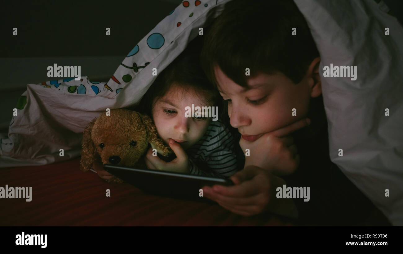 Brother and sister lying on the bed looking at the tablet in the dark Stock Photo