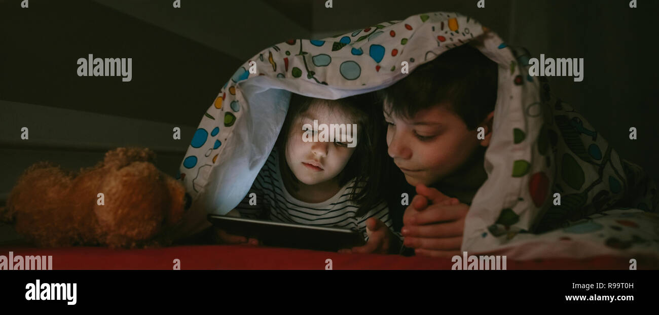 Brother and sister lying on the bed looking at the tablet in the dark Stock Photo