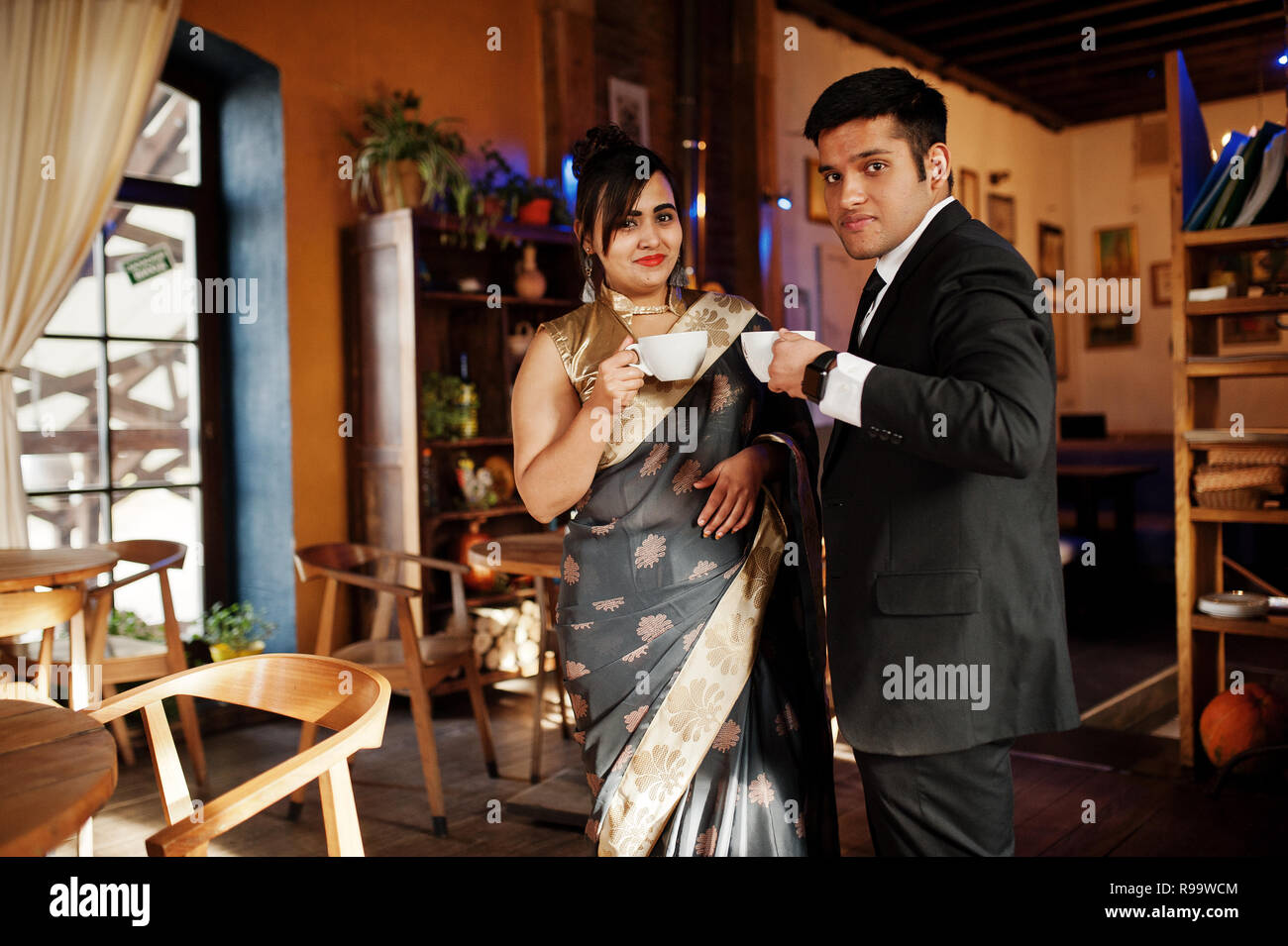 Elegant and fashionable indian friends couple of woman in saree and man in suit sitting on cafe and drinking tea. Stock Photo