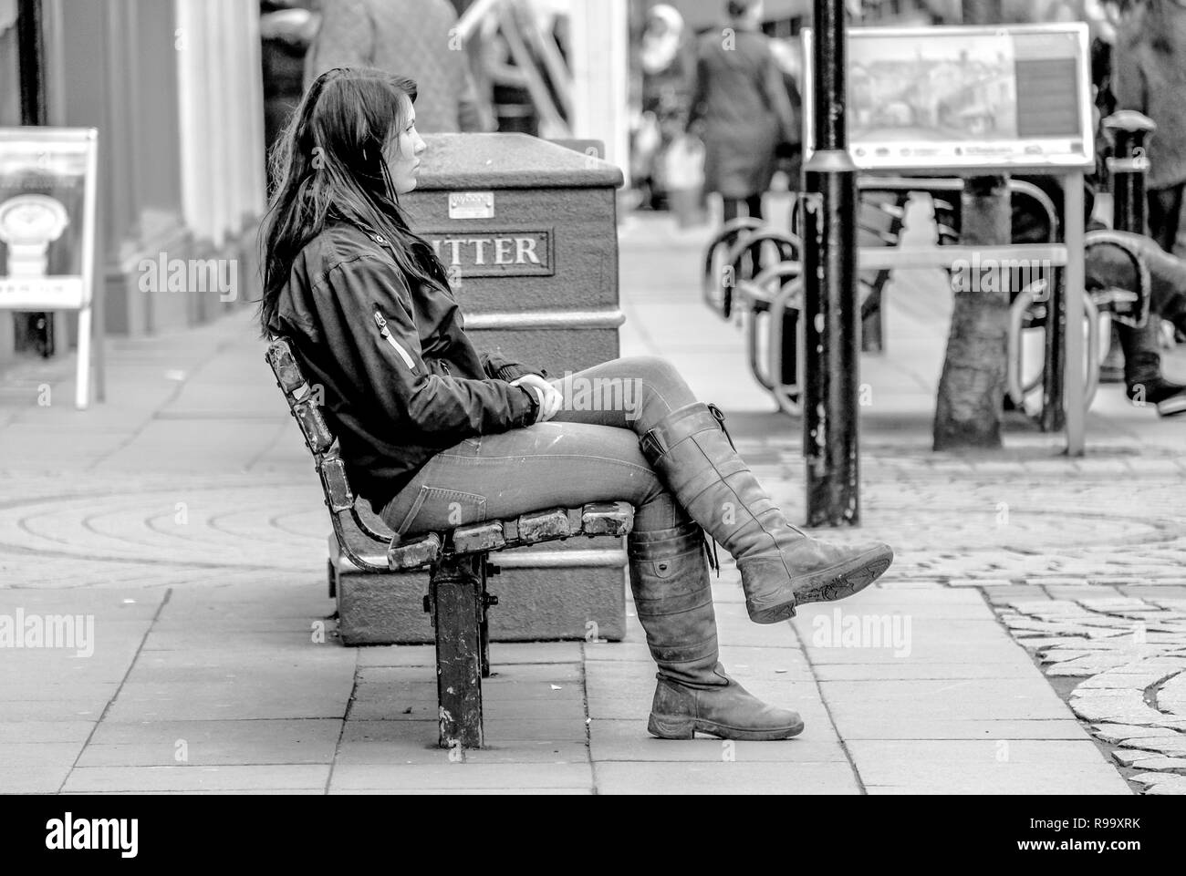 A young woman with long hair in leather jacket, boots and jeans waits on a bench within a pedestrianised urban town centre shopping area Stock Photo