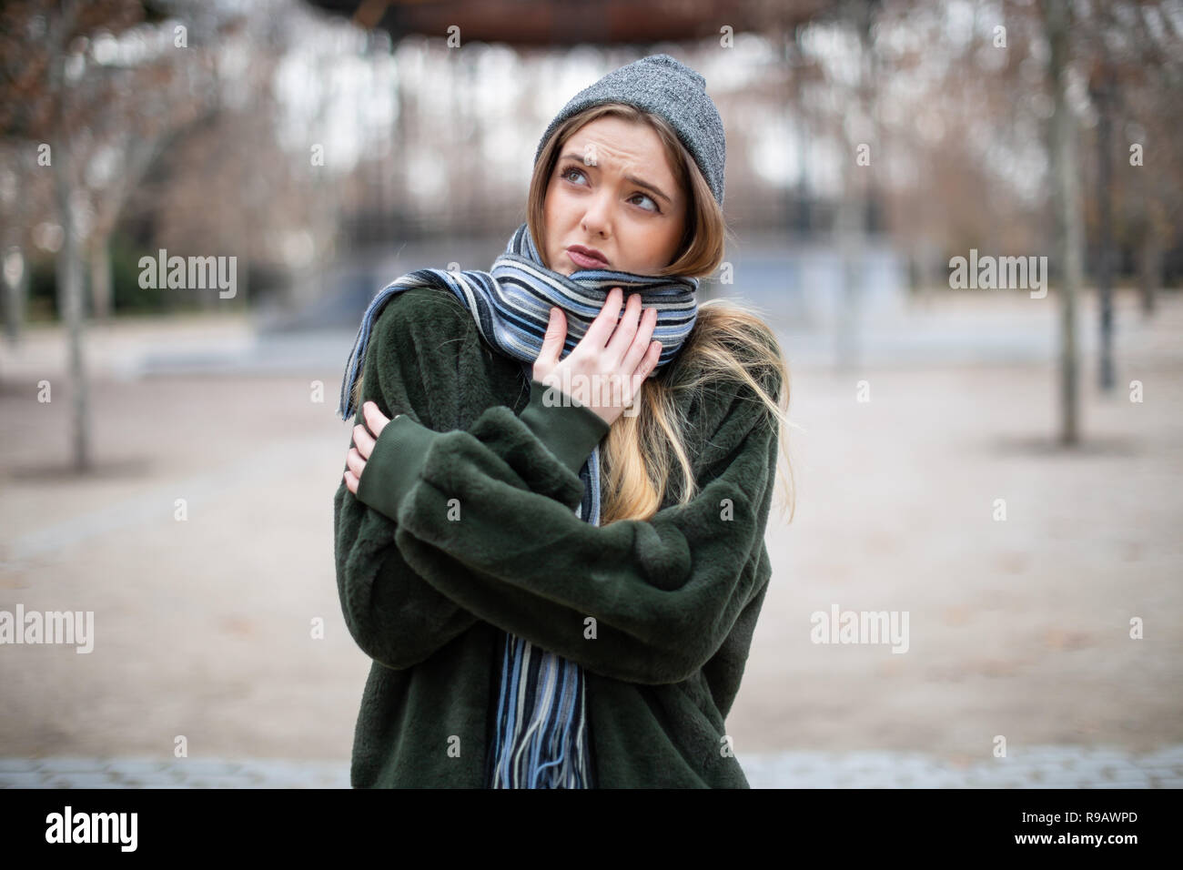Pretty young female in warm clothes wrapping in scarf and looking away while standing on blurred background of autumn park on really cold day Stock Photo