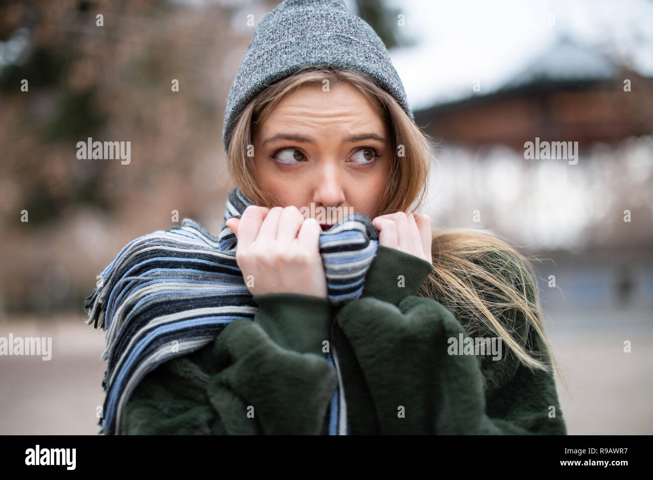 Pretty young female in warm clothes wrapping in scarf and looking away while standing on blurred background of autumn park on really cold day Stock Photo