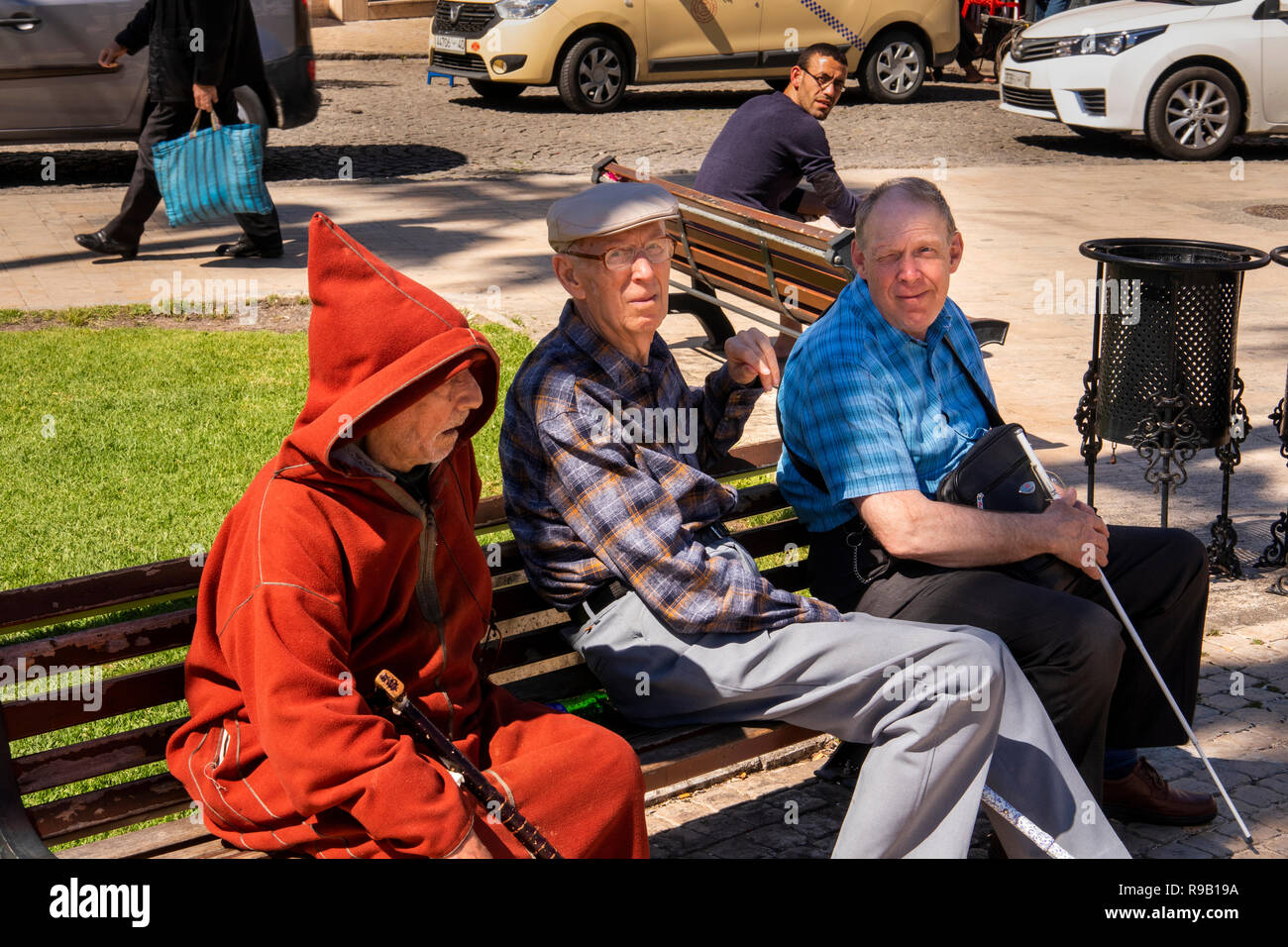 Morocco, Tangier, Place 9 Avril, Grand Socco public square, tourists sat on bench beside man in djellaba coat Stock Photo