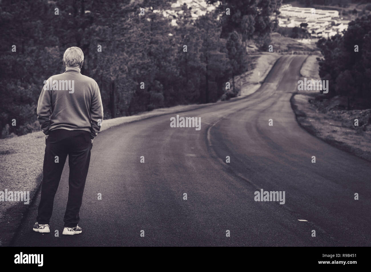 Active senior man standing on lonely road between mountains in black and white. Older man of back standing on lonely highway Stock Photo