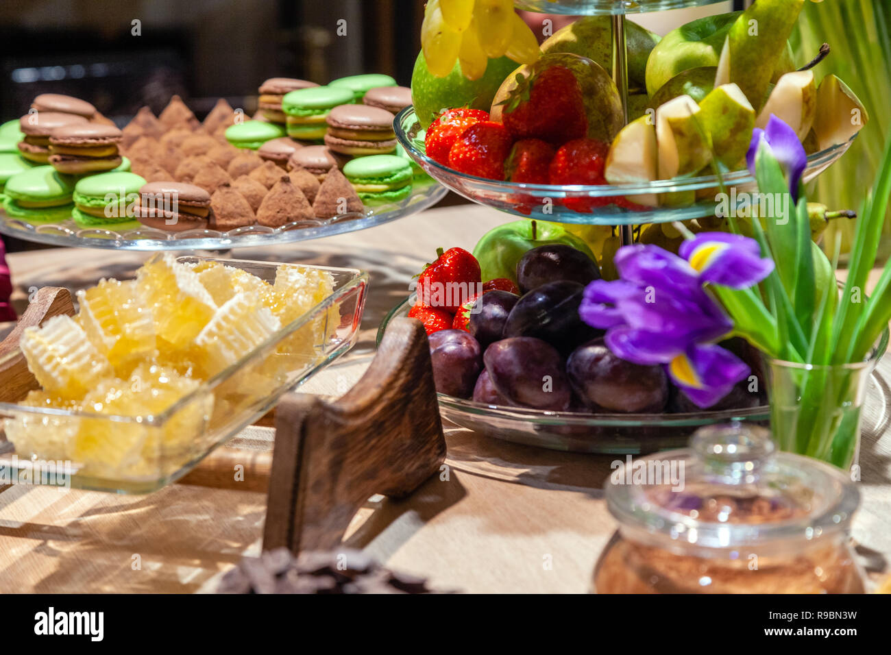 Closeup of fresh honeycombs cut into cubes in transparent square glass bowl at buffet, cinnamon sticks, fruits, macaroons, truffles, nuts, candles. Co Stock Photo