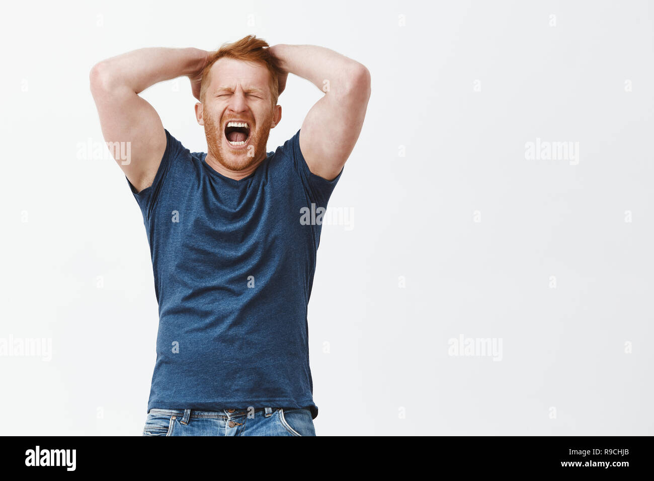 Indoor shot of redhead man feeling distress and painful emotions, holding hands on head, shouting or yelling with closed eyes, being hearbroken or losing huge money, standing depressed over grey wall Stock Photo