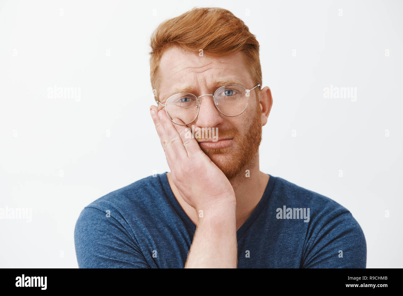 Headshot of upset and gloomy redhead guy with beard in transparent glasses, frowning holding palm on cheek gazing with regret and disappointment, losing chance over gray background Stock Photo