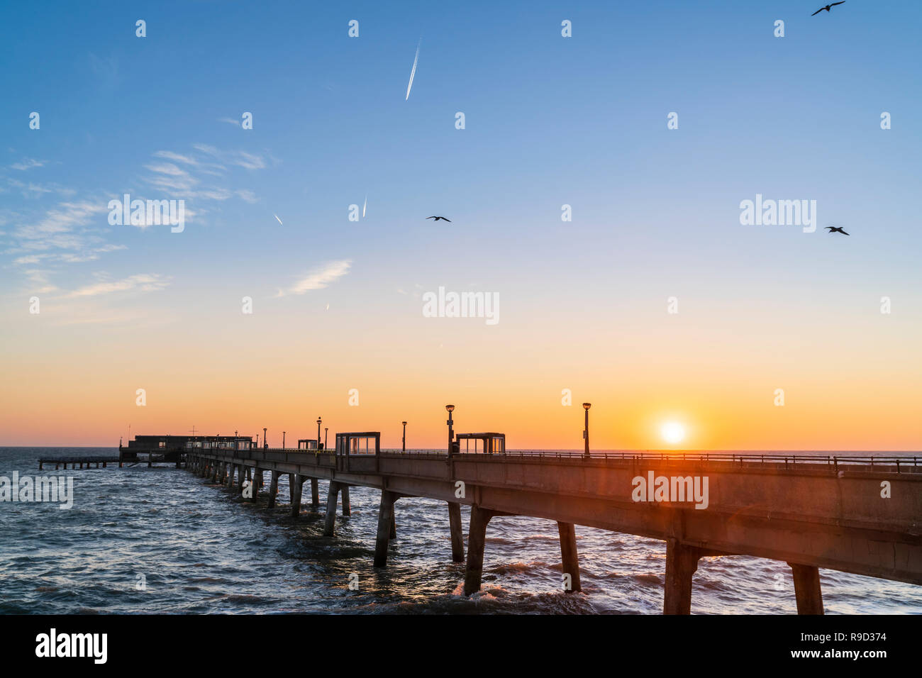 Sunrise in clear blue and yellow sky over the English Channel and Deal pier on the kent Coast. Choppy, rough sea crashing on to shingle beach. Stock Photo