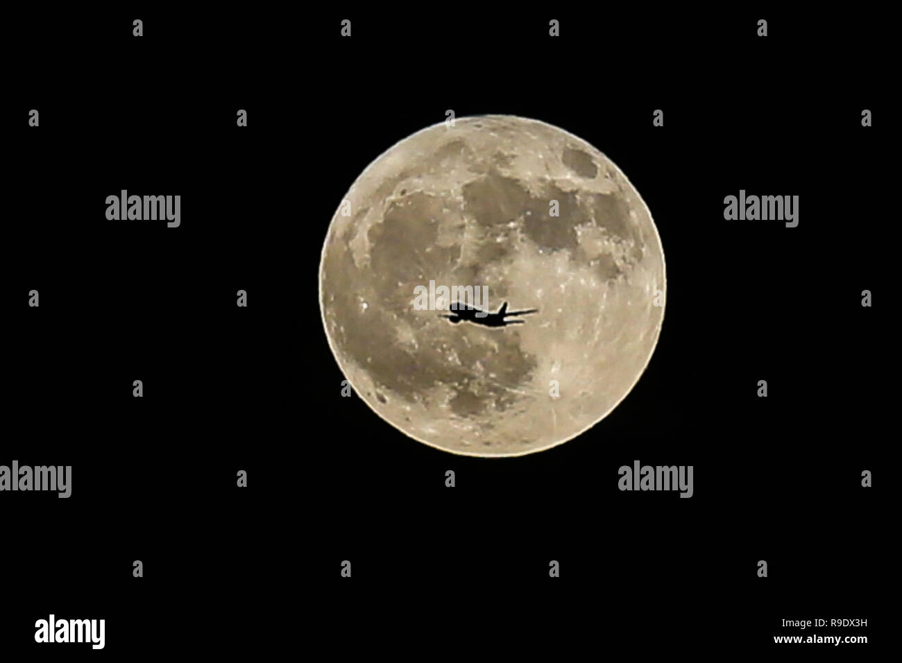 An airplane flies in front of Winter Solstice Moon over London. December's full moon has earned the nickname of the Cold Moon which falls shortly after Friday's winter solstice and the peak of the Ursid Meteor Shower - the activity that begins annually around December 17 and runs for a week plus, until the 25th or 26th. Stock Photo
