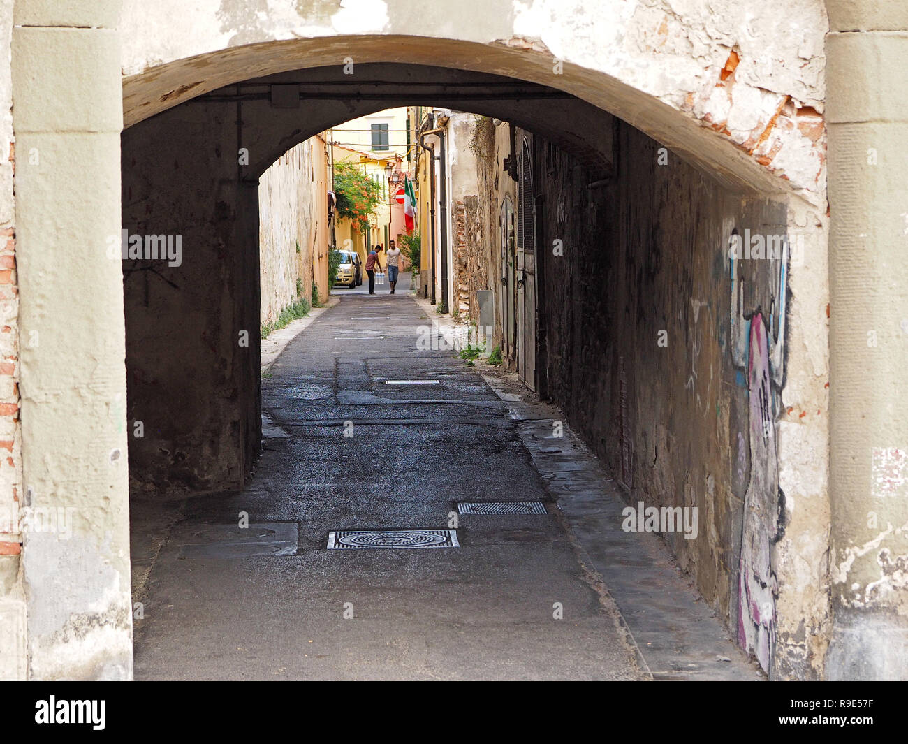two men carry pack of bottled water below shuttered windows in typical narrow street with manhole covers and graffiti under archway in Pisa, Italy Stock Photo