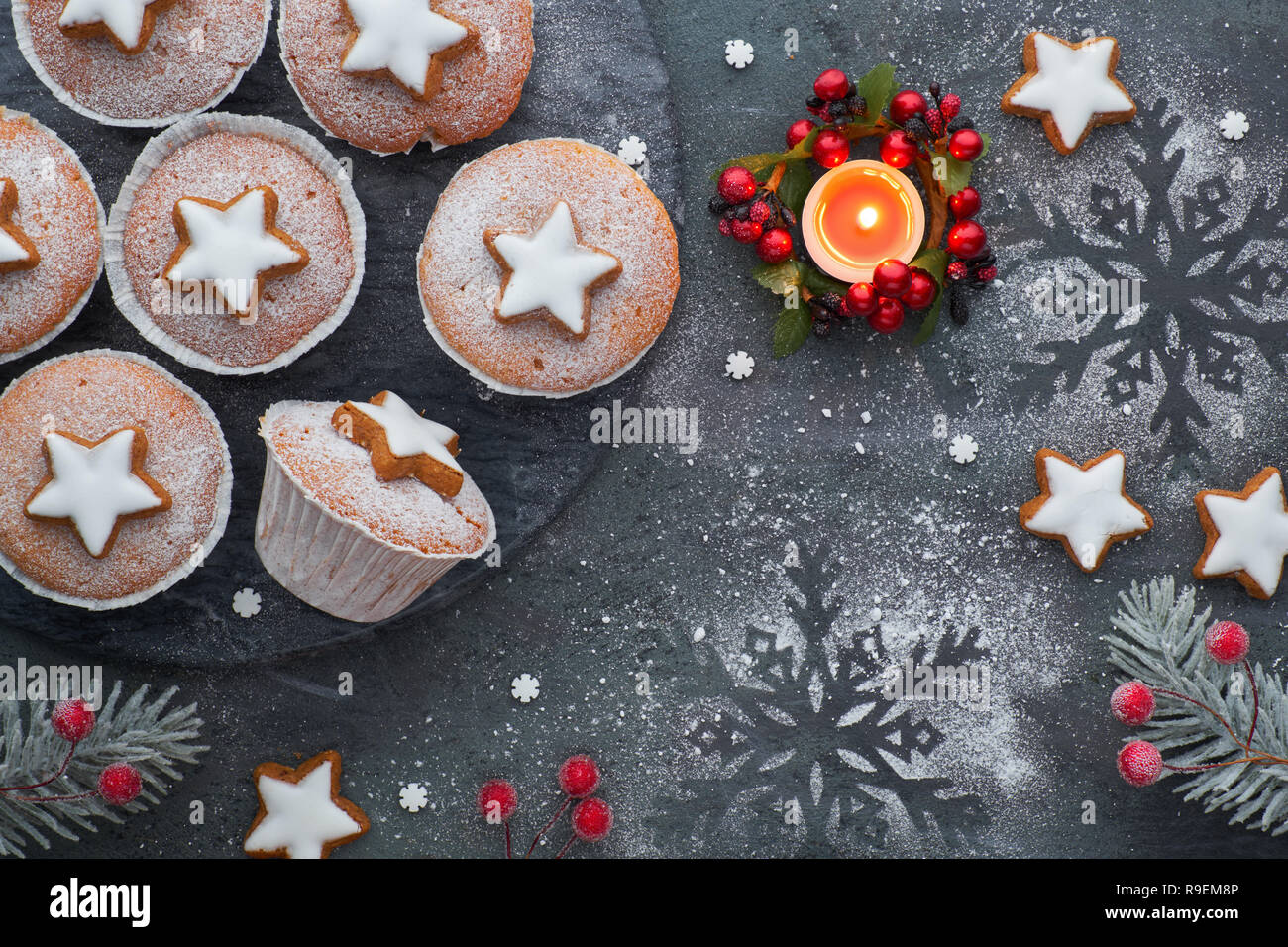 Top view of the wooden board  with sugar-sprinkled muffins, fondant icing and Christmas star cookies on dark background with candle Stock Photo