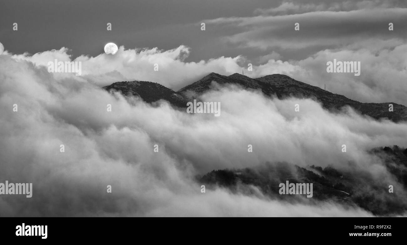 Amazing Full moon started to rise above Troodos mountains covered in evening fogy clouds. Stock Photo