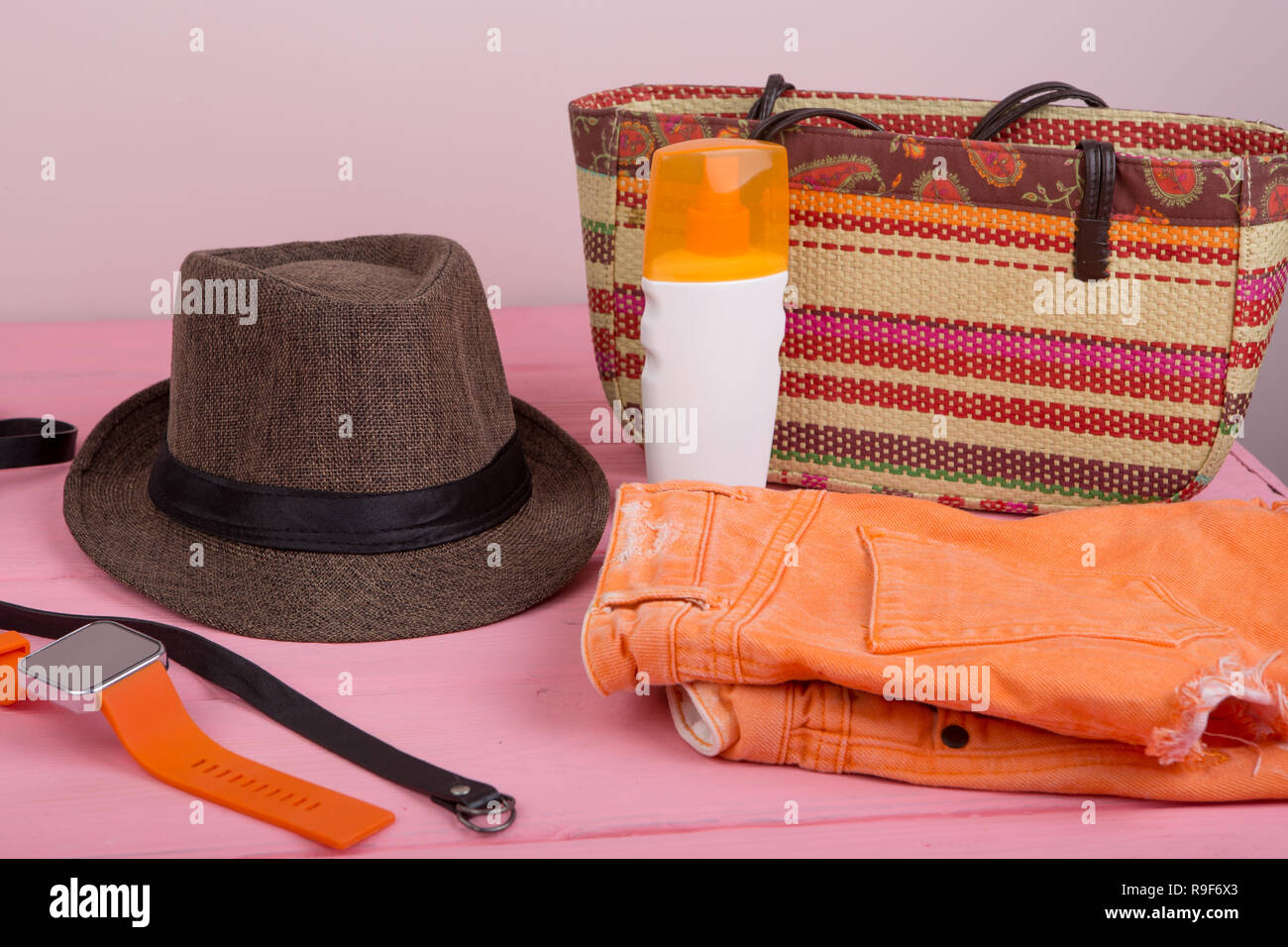 Summer accessories - straw beach bag, sun hat, belt, orange watch, denim shorts, suntan lotion on pink wooden table Stock Photo