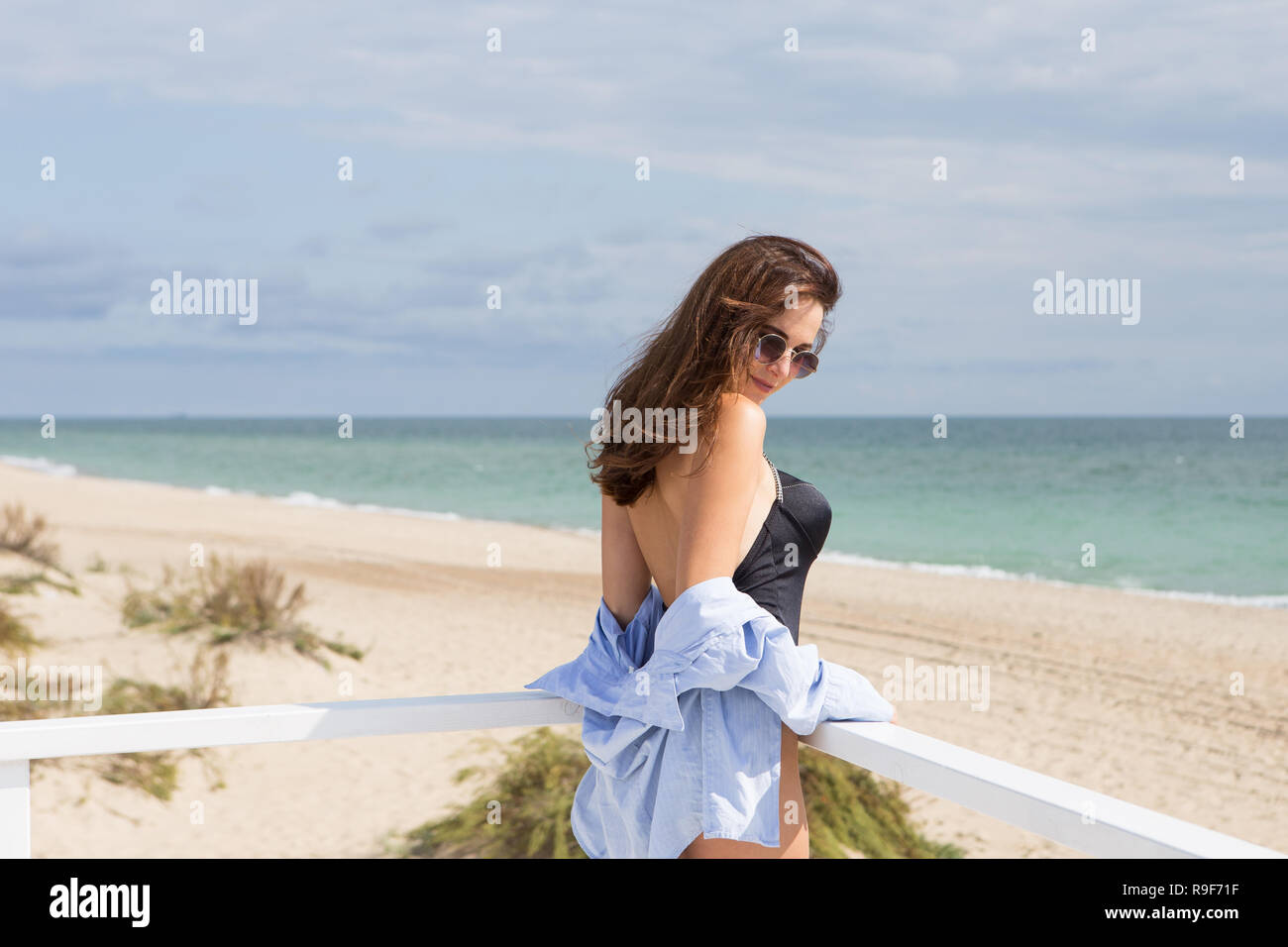 Portrait of a young sexy girl standing on a background of beach, sand and sea. She wearing black swimwear, blue shirt and sunglasses Stock Photo