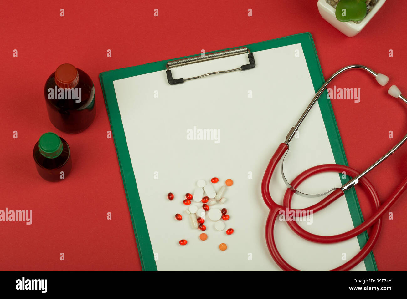 Doctor workplace - red stethoscope, pills, medical bottles and empty green clipboard on red paper background Stock Photo