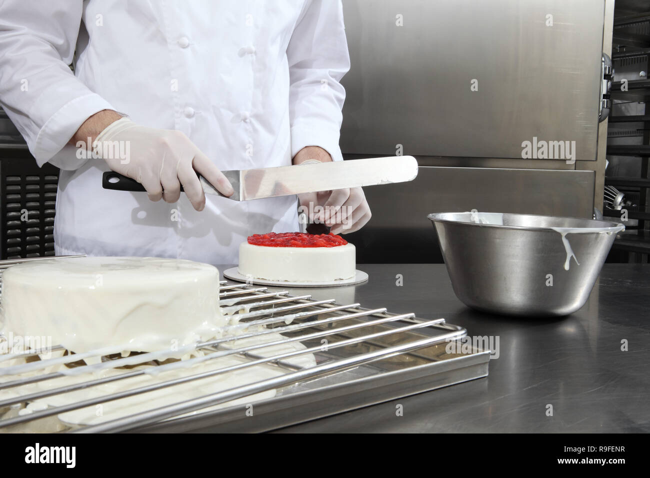 hands pastry chef prepares a cake, cover with icing and decorate with strawberries, works on a stainless steel industrial kitchen work top Stock Photo