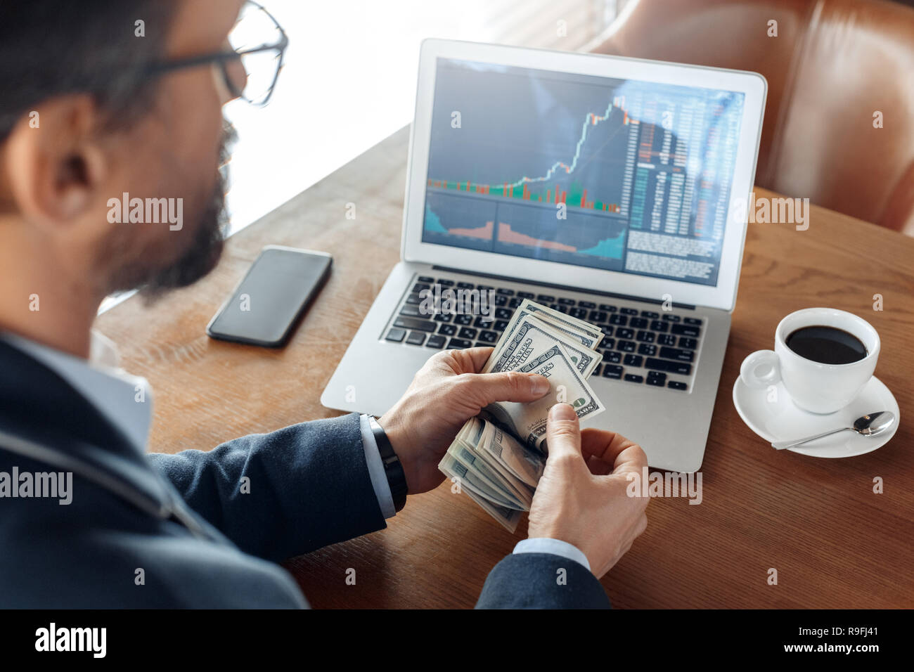 Mature man trader sitting at table at cafe with cup of coffee looking at  laptop with trading chart counting dollar banknotes back view close-up  Stock Photo - Alamy