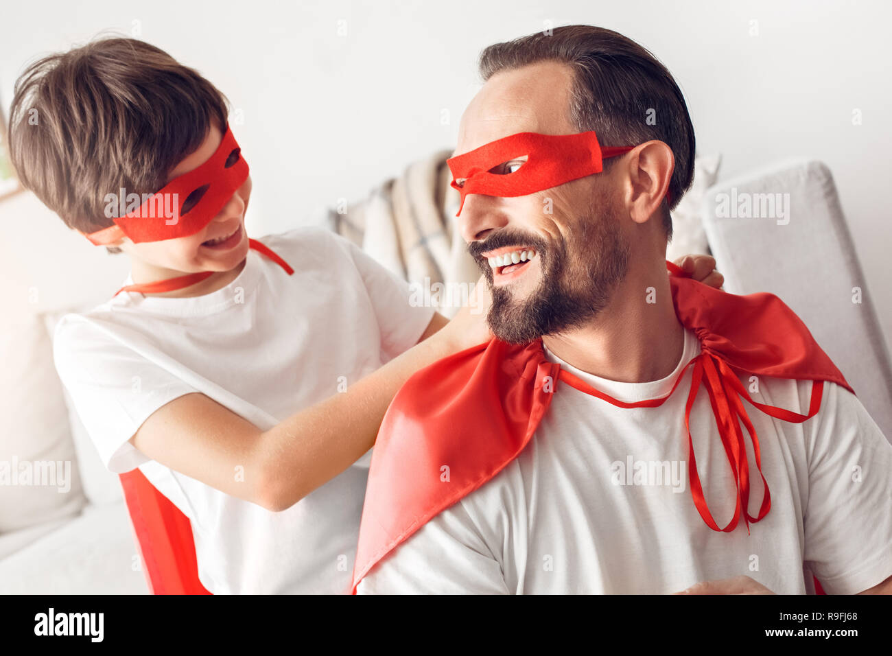 Father and little son wearing superheroe costumes together at home boy helping man to tie mask smiling cheerful close-up Stock Photo