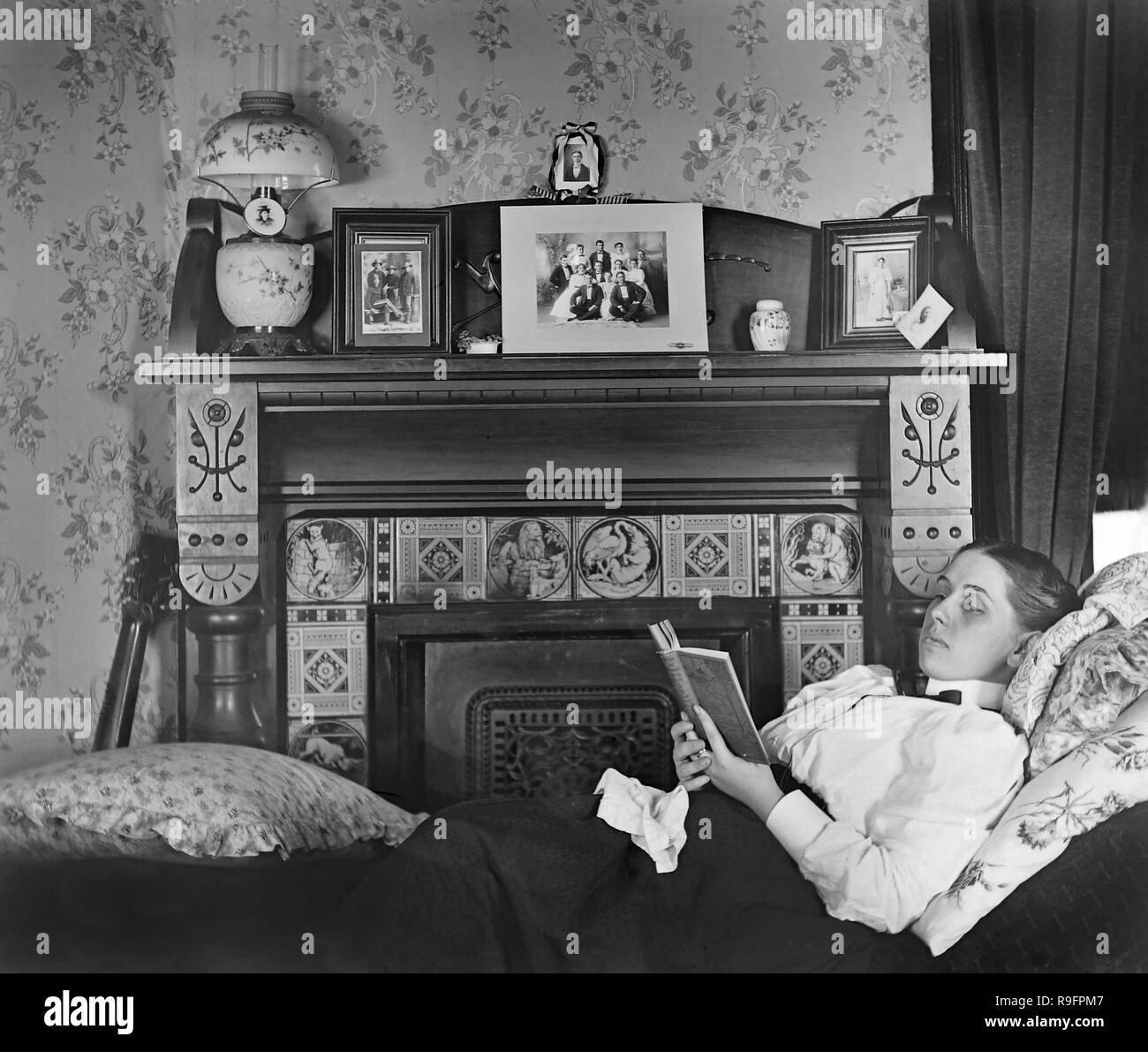 A young woman reads on a fainting couch before an ornately decorated hearth and mantle in an early 20th century parlor, ca. 1905. Stock Photo