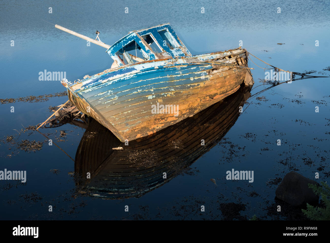 Wreck of a fishing boat on the wild atlantic coast of Ireland.  Faded paint blue ship in lying sideways with perfect reflection in low tide waters. Stock Photo