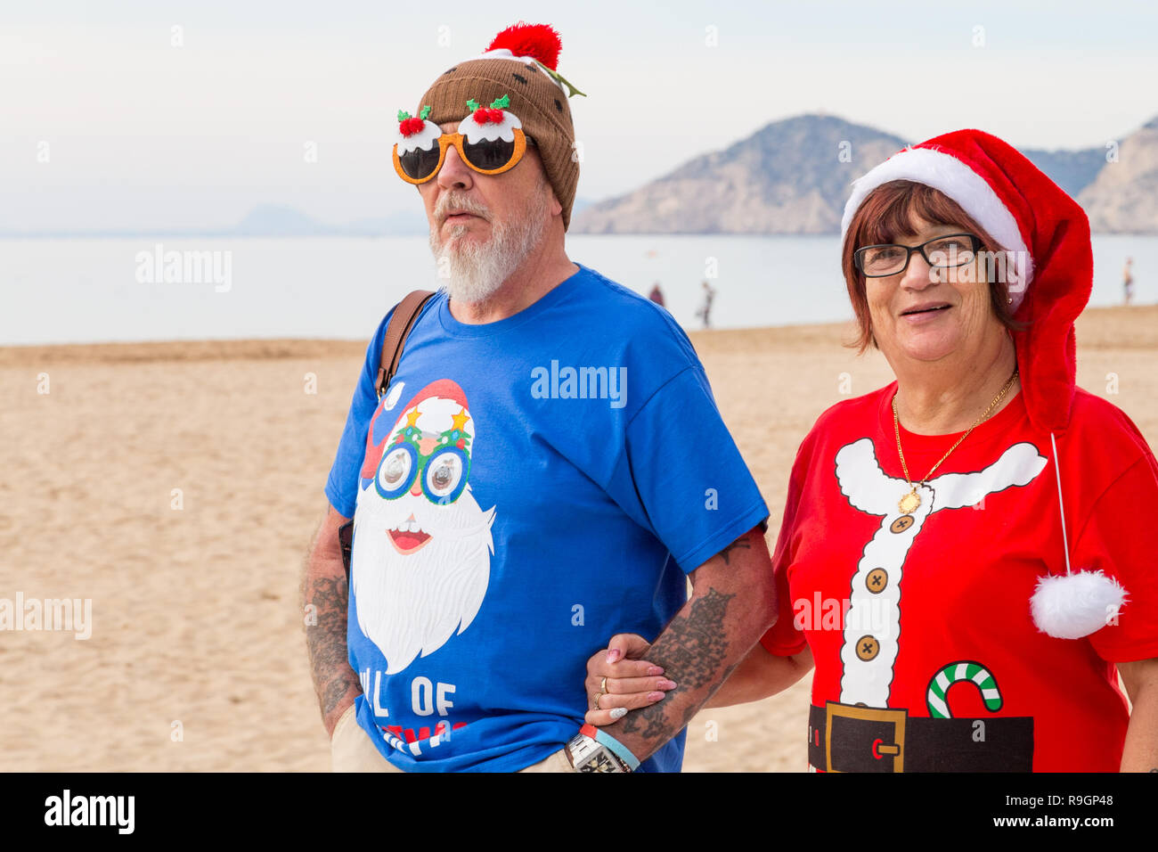 Benidorm, Costa Blanca, Spain, 25th December 2018. British tourists dress for the occasion on Christmas Day in this favourite getaway destination for Brits escaping the cold weather at home. Temperatures will be in the mid to high 20's Celsius today in this mediterranean hotspot. Middle aged couple wearing Christmas Jumpers walking outside on the sea front promenade on Levante Beach with christmas pudding comedy glasses, Stock Photo