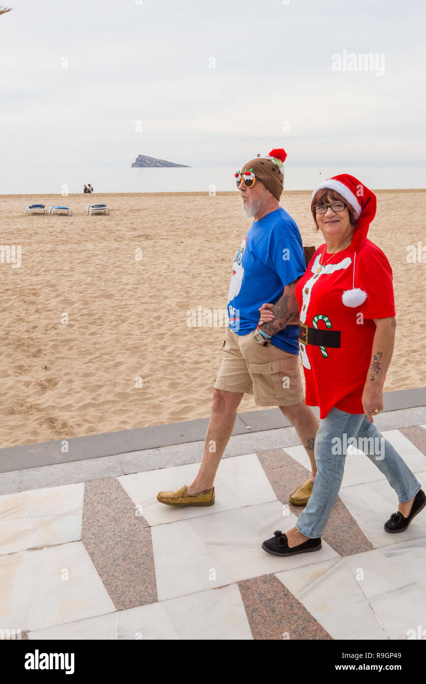 Benidorm, Costa Blanca, Spain, 25th December 2018. British tourists dress for the occasion on Christmas Day in this favourite getaway destination for Brits escaping the cold weather at home. Temperatures will be in the mid to high 20's Celsius today in this mediterranean hotspot. Middle aged couple wearing Christmas Jumpers walking outside on the sea front promenade on Levante Beach with christmas pudding comedy glasses, Stock Photo