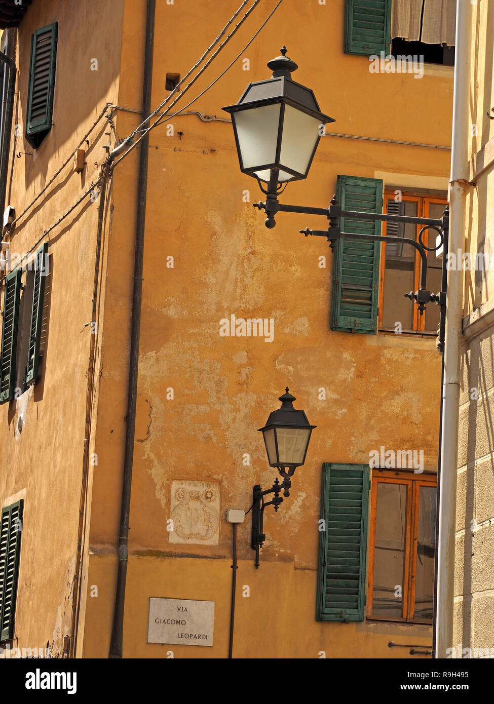 luminous lantern style street lights on iron brackets shine in sunlight against typical ochre buildings & green shuttered windows in Pisa, Italy Stock Photo