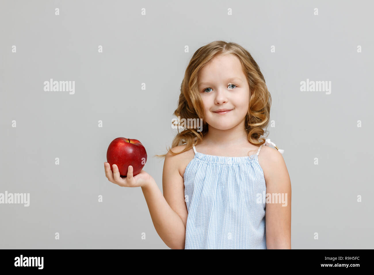 Portrait of a happy smiling little blonde girl on a gray background. The child is holding a red apple Stock Photo