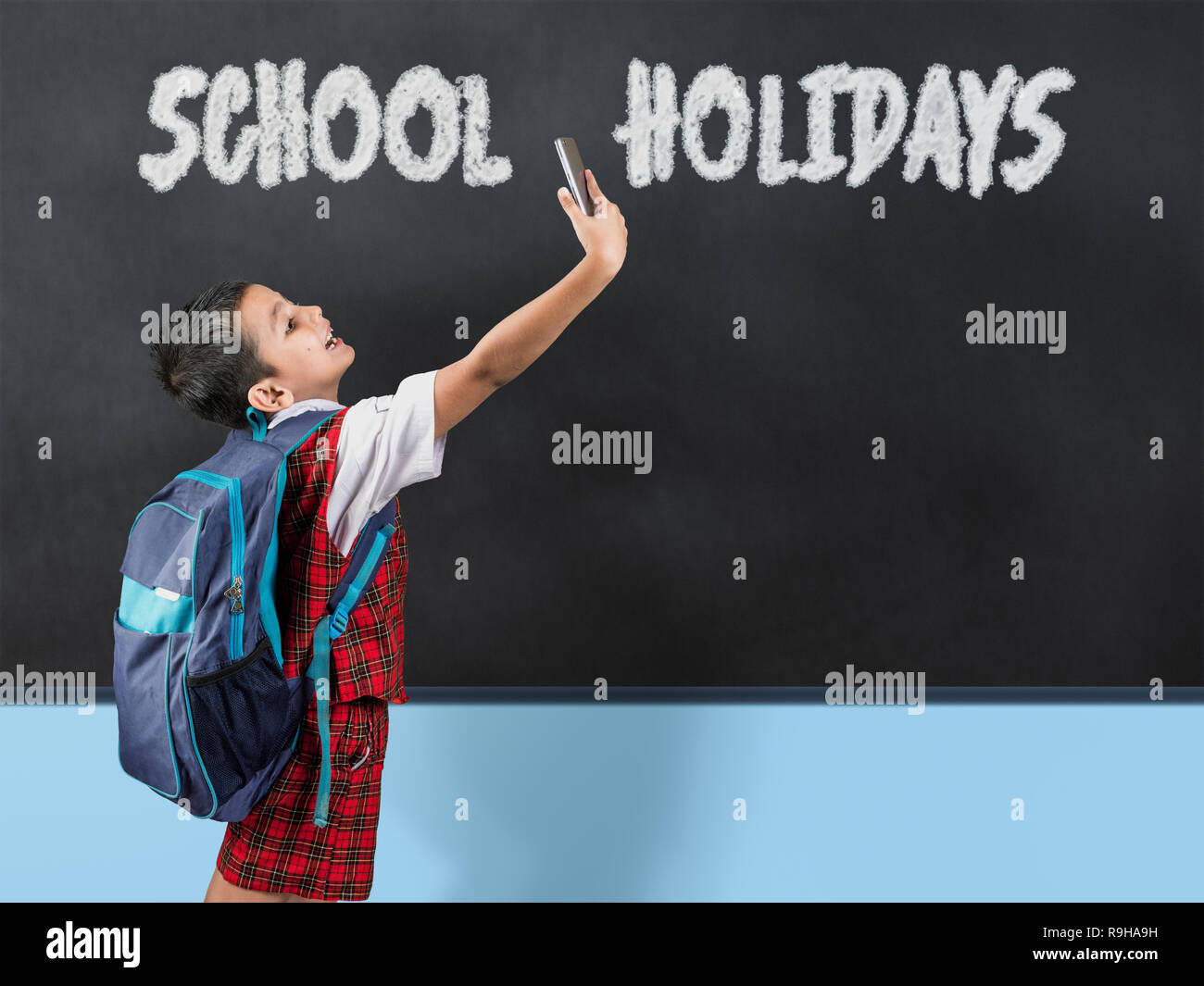 Young asian school boy wearing red uniform taking selfie with mobile phone infront of blackboard and written text 'school holidays' Stock Photo