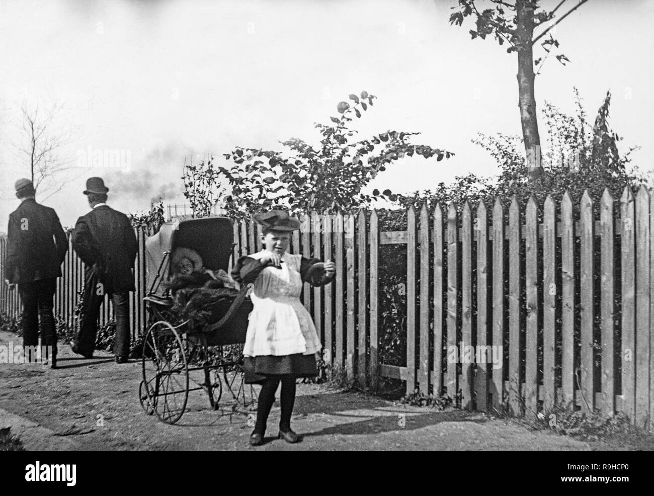 A late Victorian black and white photograph showing a very young girl with a pram with a bay in it, on a street in England, while two men walk away behind the pram. The pram has large iron wheels, and all the figures show good examples of fashions of the era. Stock Photo