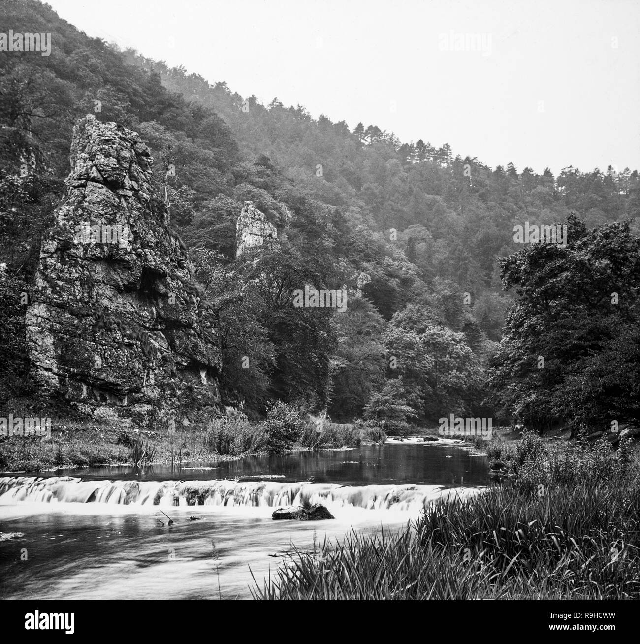 A late Victorian black and white photograph showing Dovedale, a valley in the Peak District of England. The land is now owned by the National Trust, and annually attracts a million visitors The valley was cut by the River Dove and runs for just over 3 miles (5 km) between Milldale in the north and a wooded ravine near Thorpe Cloud and Bunster Hill in the south. In the wooded ravine, a set of stepping stones cross the river, and there are two caves known as the Dove Holes. Stock Photo