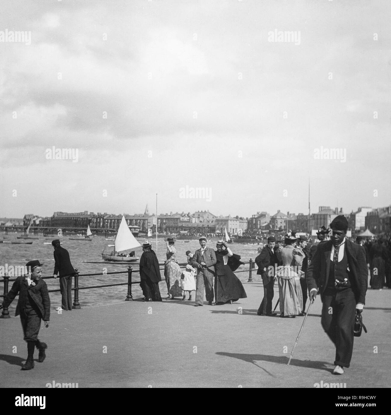 Alate Victorian black and white photograph taken in the English sea side town of Morecambe, in Lancashire, England. It shows a man of black African descent, dressed in fine clothes, and with a walking cane, walking down the promenade, whilst a young boy looks at him. There are many other people in the background showing the fashions of the day, as well as small boats in the harbour. Stock Photo