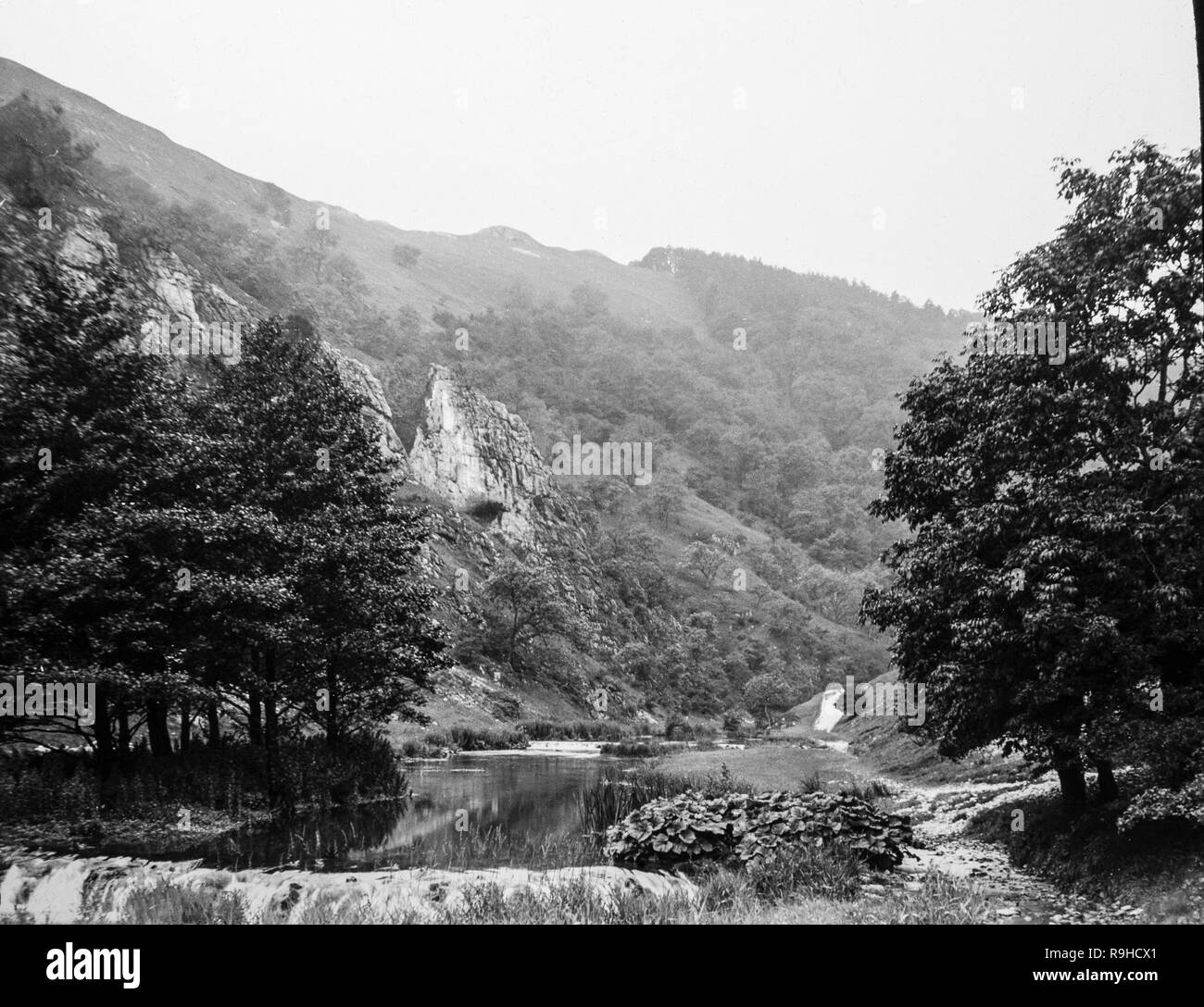 A late Victorian black and white photograph showing Dovedale, a valley in the Peak District of England. The land is now owned by the National Trust, and annually attracts a million visitors The valley was cut by the River Dove and runs for just over 3 miles (5 km) between Milldale in the north and a wooded ravine near Thorpe Cloud and Bunster Hill in the south. In the wooded ravine, a set of stepping stones cross the river, and there are two caves known as the Dove Holes. Stock Photo