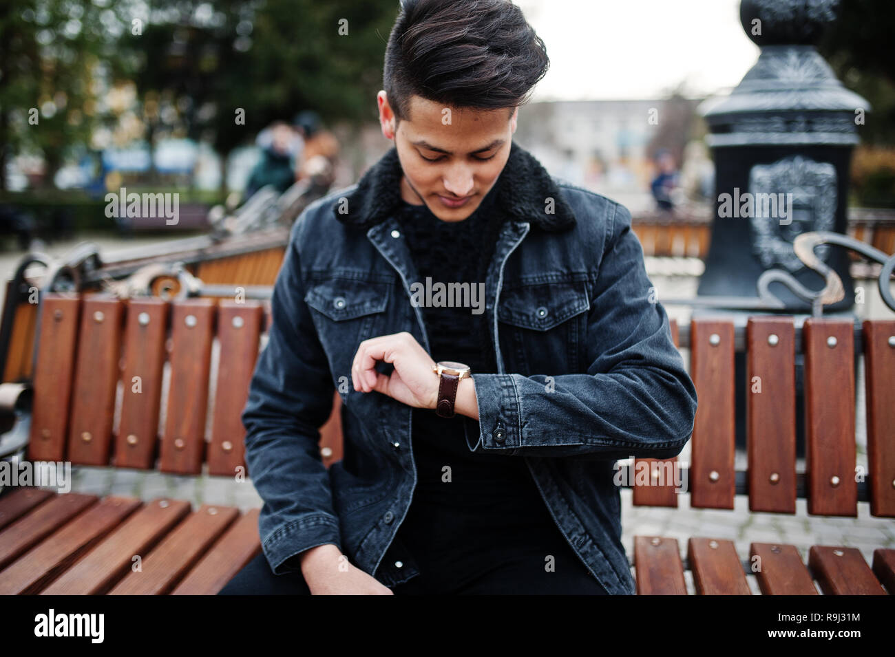 Handsome and fashionable indian man in black jeans jacket posed outdoor,  sitting on bench and looking at his watches Stock Photo - Alamy