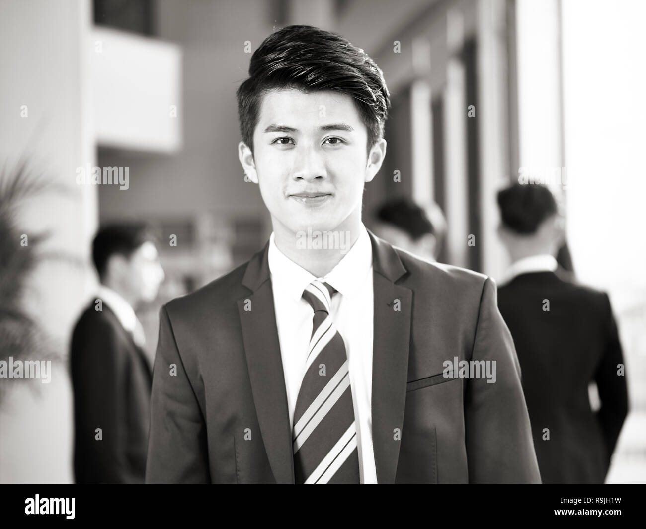 portrait of a young asian business man, pointing and looking at camera smiling, colleagues chatting in background, black and white. Stock Photo